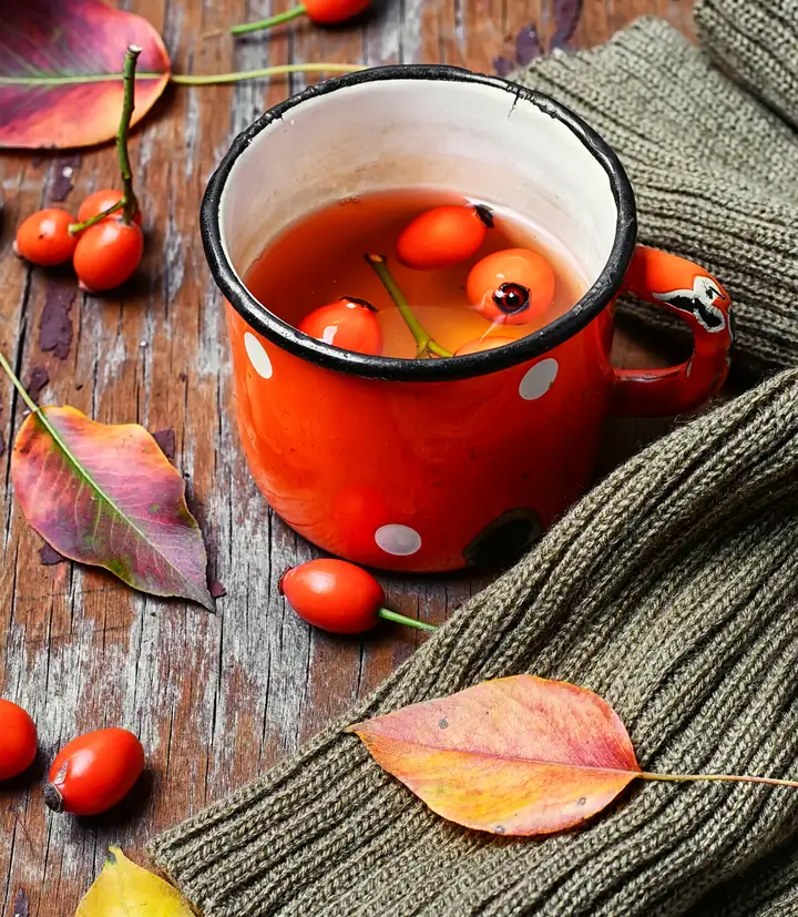 A red enamel mug of rose hip tea on a weathered table with autumn leaves and a knit sweater